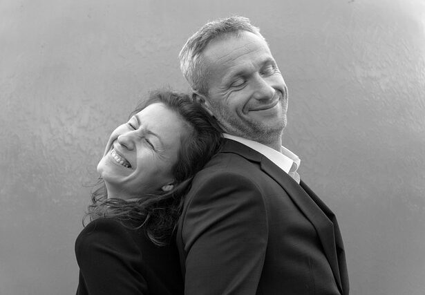 VSP Erding Ein Mann und eine Frau in Business-Kleidung stehen Rücken an Rücken, beide lächeln mit geschlossenen Augen vor einem schlichten Hintergrund. Dieses Schwarz-Weiß-Foto fängt Ehepaare aus dem Landkreis Erding perfekt in einem Moment der Harmonie ein. Rechtsanwalt Erding