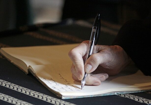 VSP Erding Nahaufnahme der Hand einer Person, die einen Stift hält und in einem aufgeschlagenen Notizbuch über Ansprüche Erbrecht schreibt. Das Notizbuch liegt auf einer dunklen Oberfläche mit einem gemusterten Stoff, während der handgeschriebene Text auf der Seite teilweise sichtbar ist. Rechtsanwalt Erding